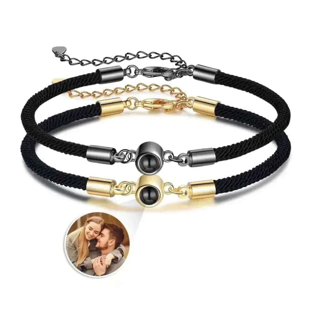 To armbånd i sort og guldfarvet nylonsnor med et rhodineret charm, der gemmer et billede bag en linse.