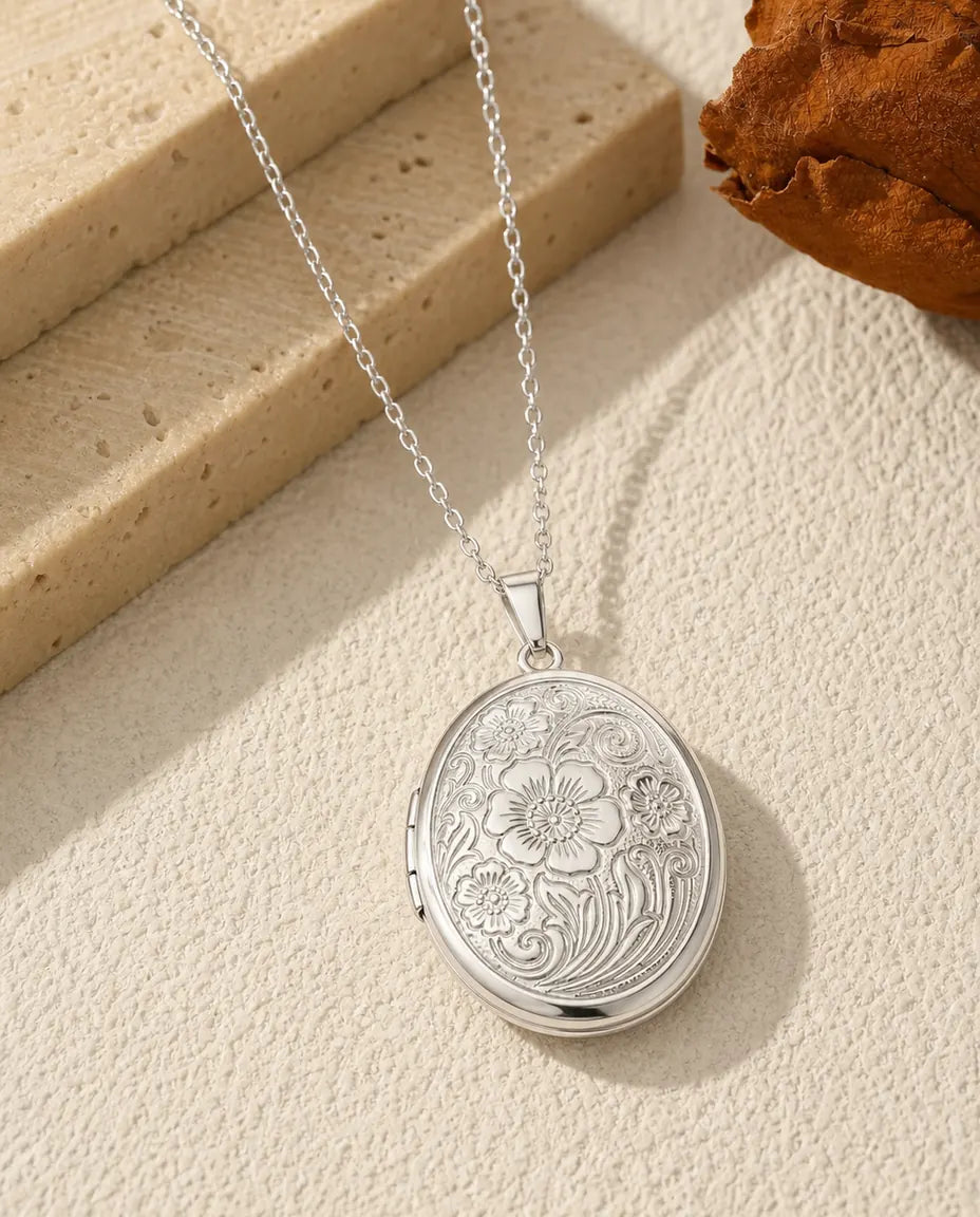 Aflang oval medaljon i rustfrit stål med markblomster i relief på forsiden og to ovale billedrammer indeni.
