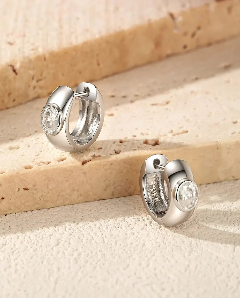 To brede creoler i 925 sterling sølv med en oval moissanite foran ligger på et lyst trin af travertin i varmt sidelys.