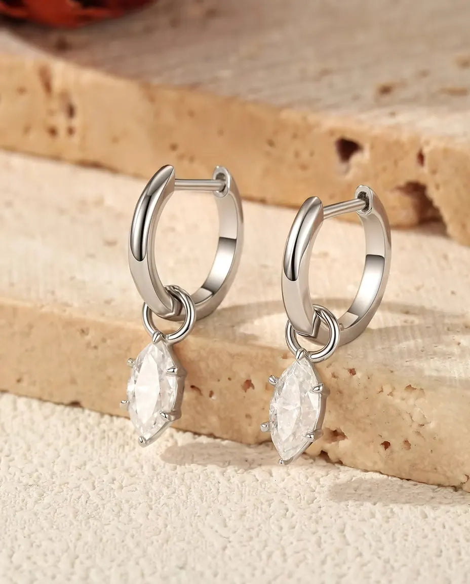 Et par små creoler i 925 sterling sølv med hængende marquise moissanite står på et lyst trin af travertin ved en tørret rose.