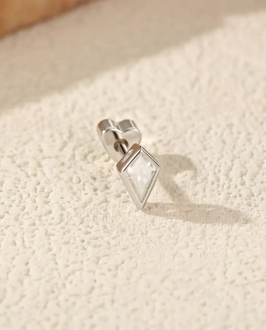 En kite-slebet moissanite ørestik i 925 sterling sølv med hjerteformet lås ligger på skrå på råhvid puds i varmt sidelys.