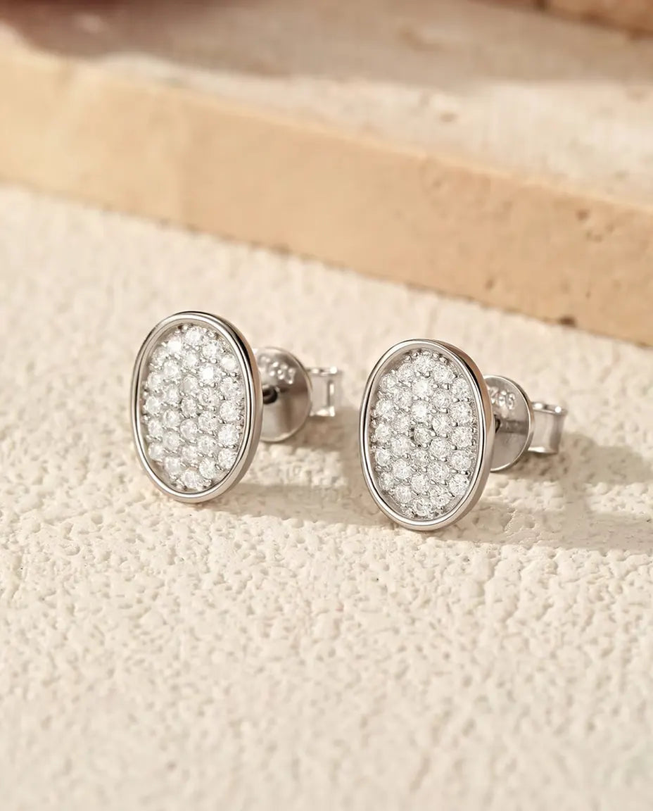 Et par ovale ørestikker i 925 sterling sølv, dækket af små moissanite-sten, står side om side på råhvid puds ved travertin.