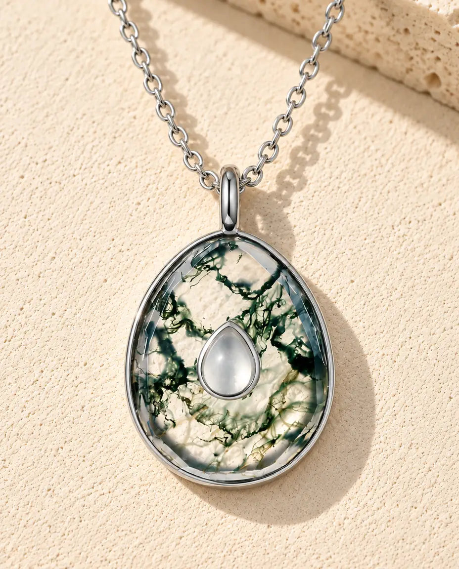 Halskæde i sølv med en ægformet, klar moss agate med grøn mos og en lille hvid dråbe i midten på beige puds foran travertin.