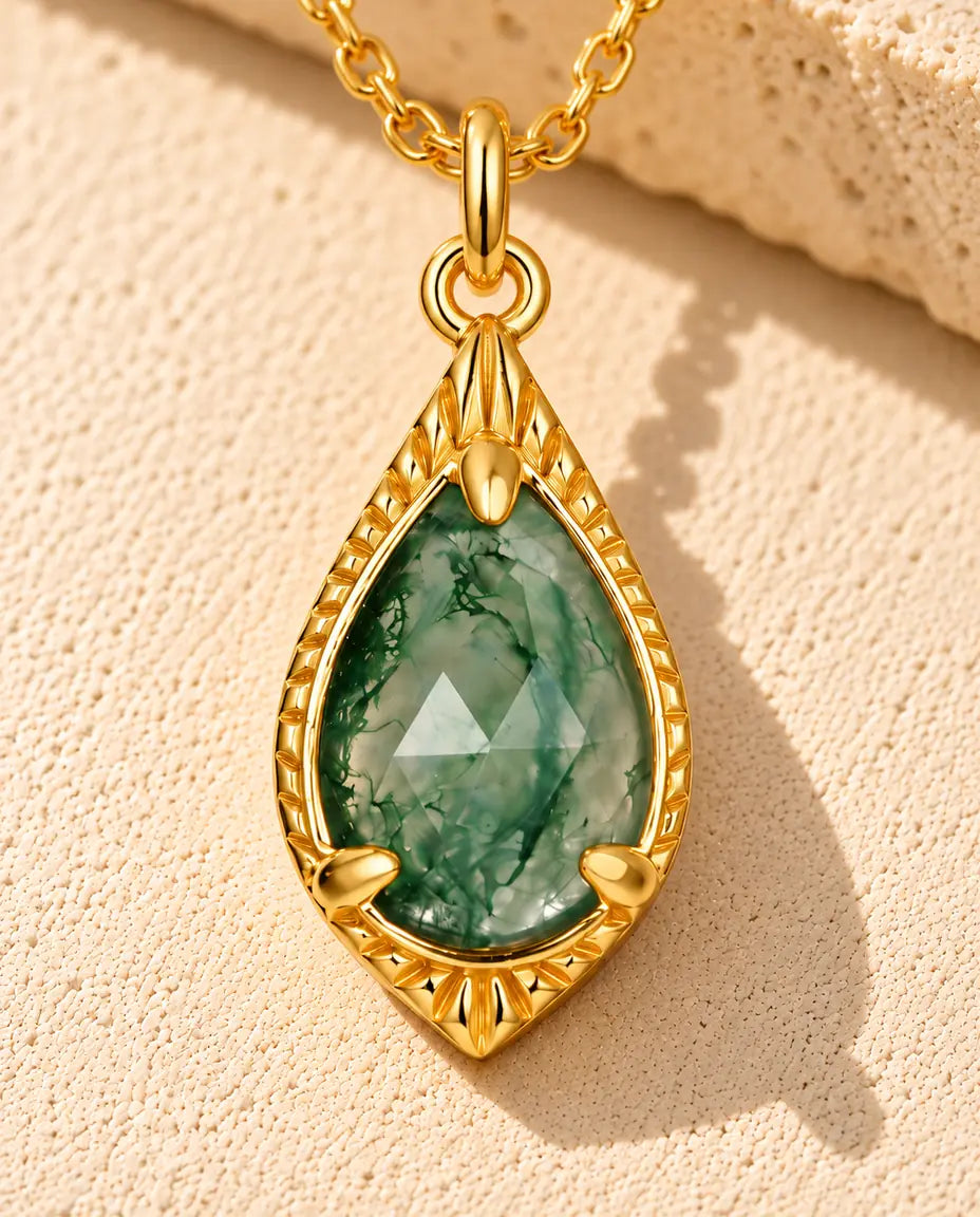 Guldbelagt halskæde med en grøn facetteret moss agate-dråbe i en guldramme med fin perlekant på beige puds foran travertin.