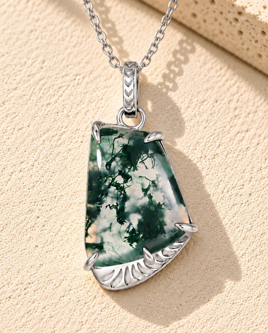 Halskæde i sølv med en klar, trapezformet moss agate med mørkegrøn mos på en fatning med fjermønster foran travertin.