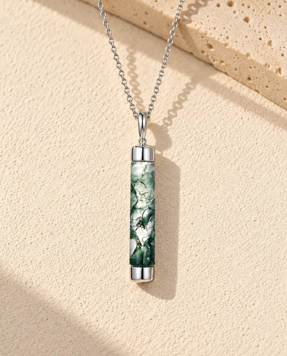 Halskæde i sølv med en klar moss agate-cylinder med grøn mos mellem to blanke sølvhætter på beige puds foran travertin.