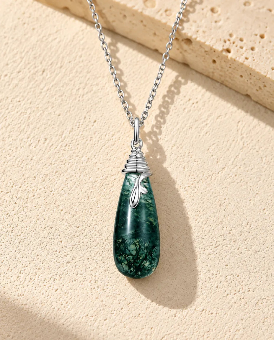 Sølvhalskæde med en lang mørkegrøn moss agate-dråbe under en hætte af snoet sølvtråd med en bladformet ranke, på beige puds.