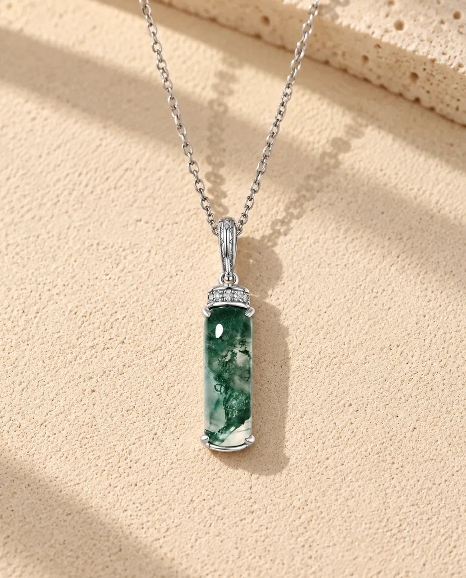 Halskæde i sølv med en mørkegrøn moss agate-stav og en krans af små hvide zirkonia foroven på beige puds foran travertin.