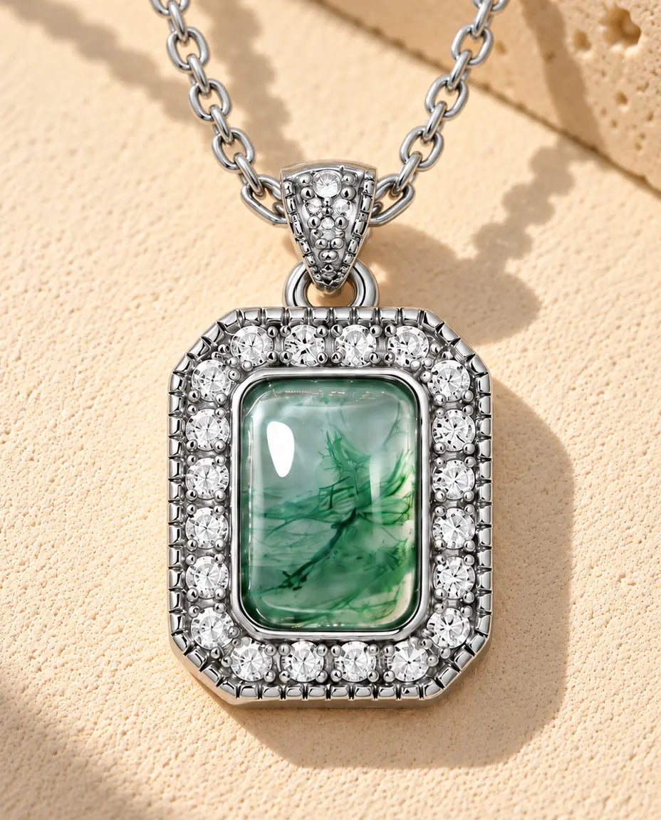 Sølvhalskæde med en lille lysegrøn moss agate i en ring af små hvide zirkonia og en perlekant på beige puds foran travertin.