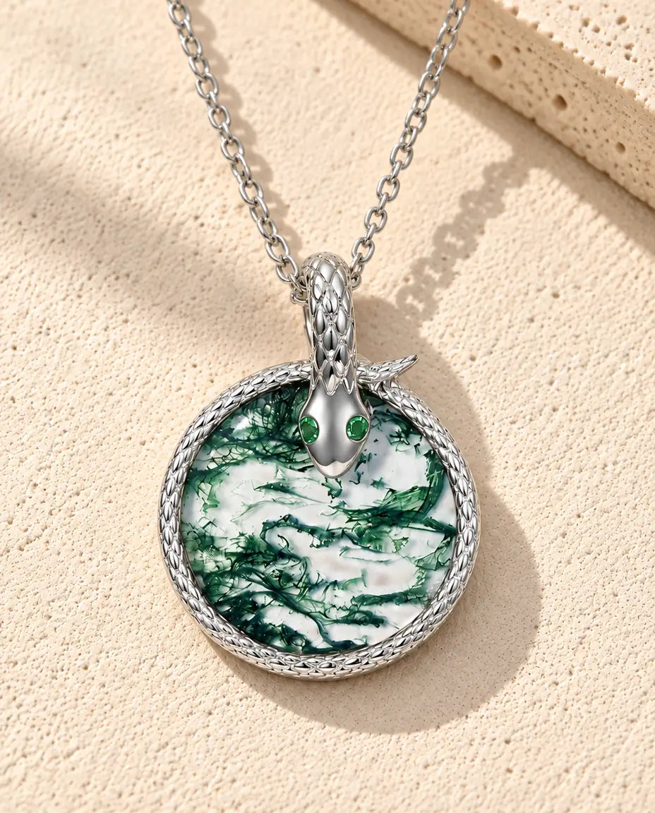 Sølvhalskæde med en lille slange med skæl om en klar, grøn rund moss agate og to grønne øjne på beige puds foran travertin.