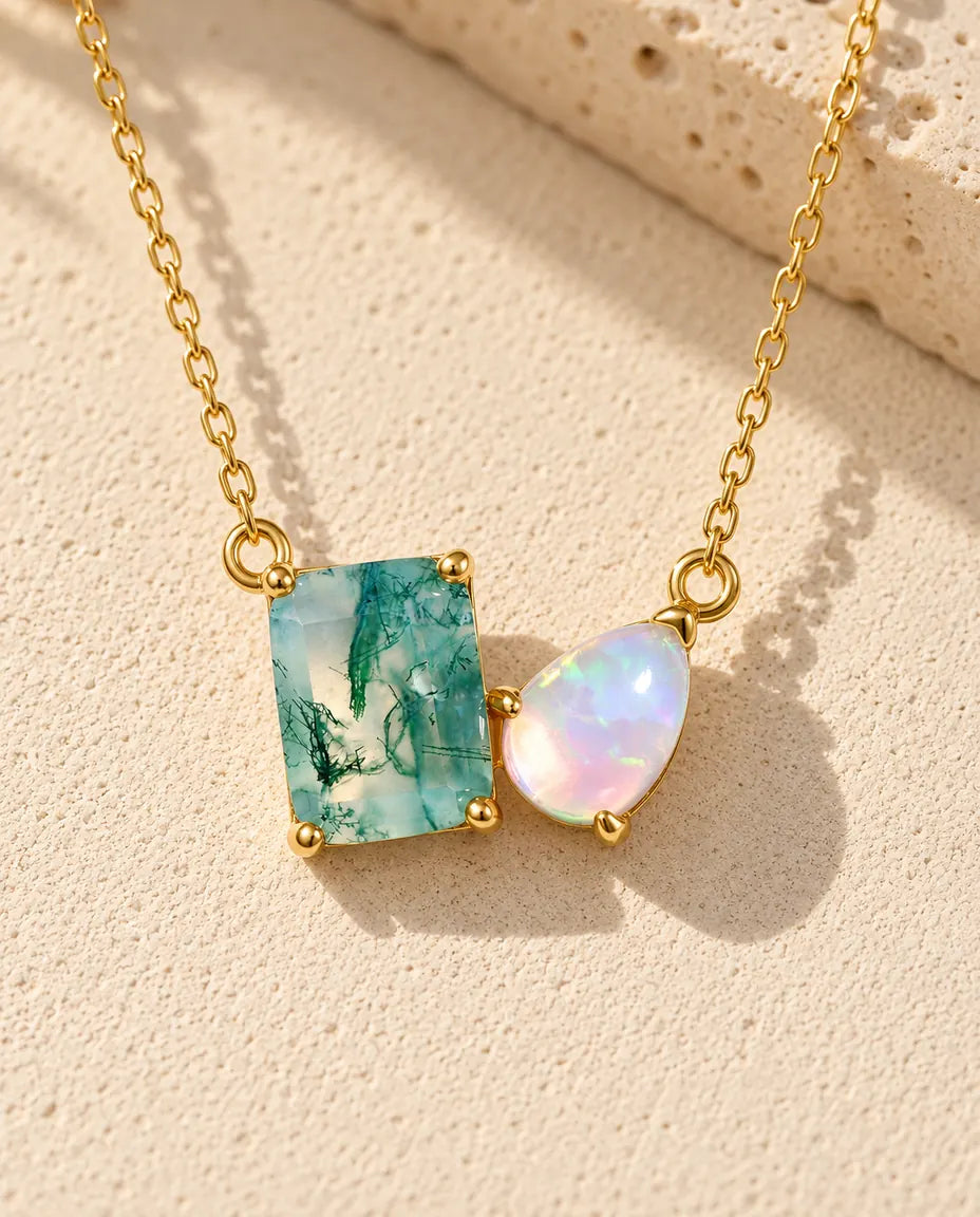 Guldbelagt halskæde med en rektangulær grøn moss agate og en dråbeformet opal på beige puds foran travertin i varmt sidelys.