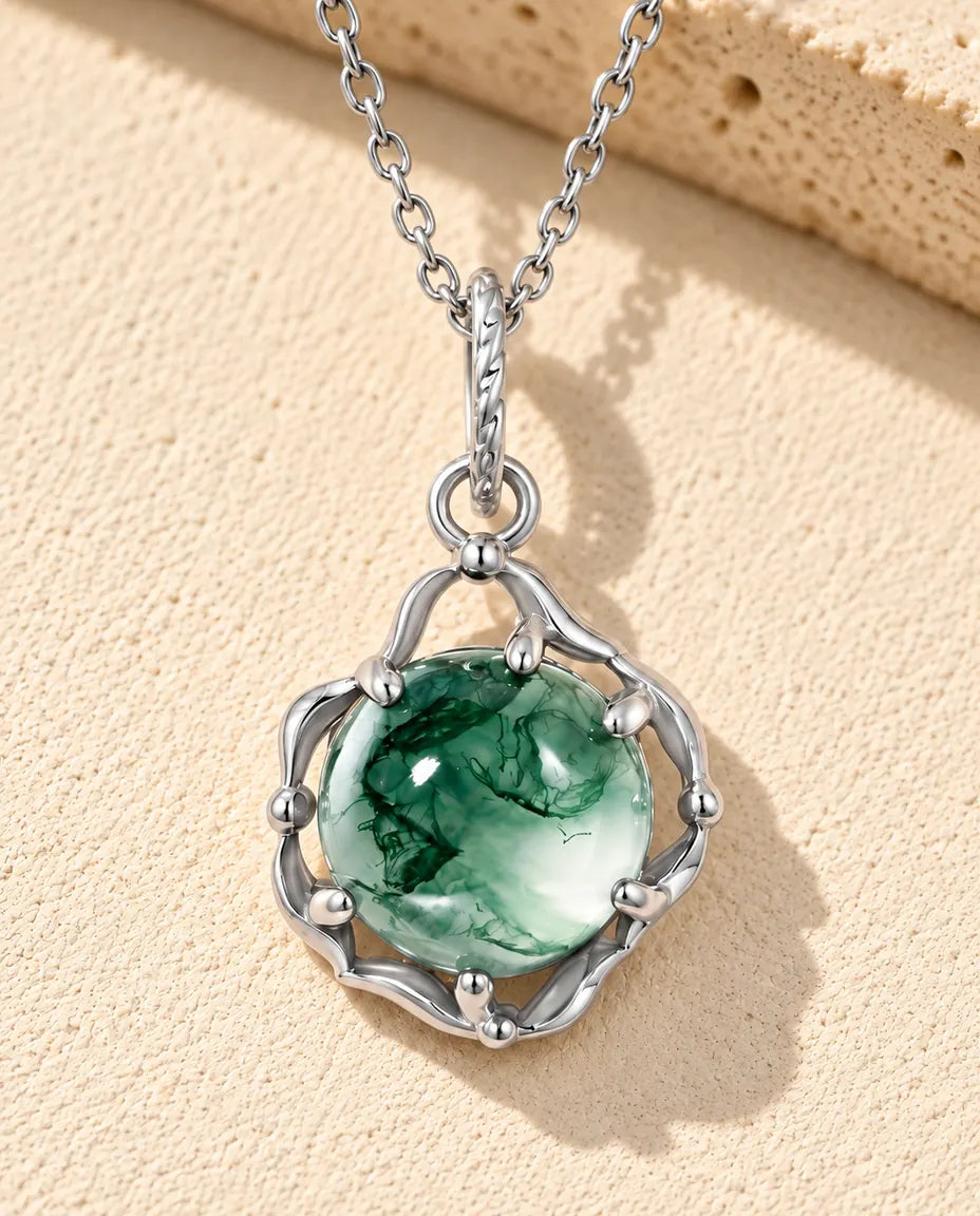 Halskæde i sølv med en grøn rund moss agate-cabochon omsluttet af bølgede sølvbånd på beige puds foran travertin.