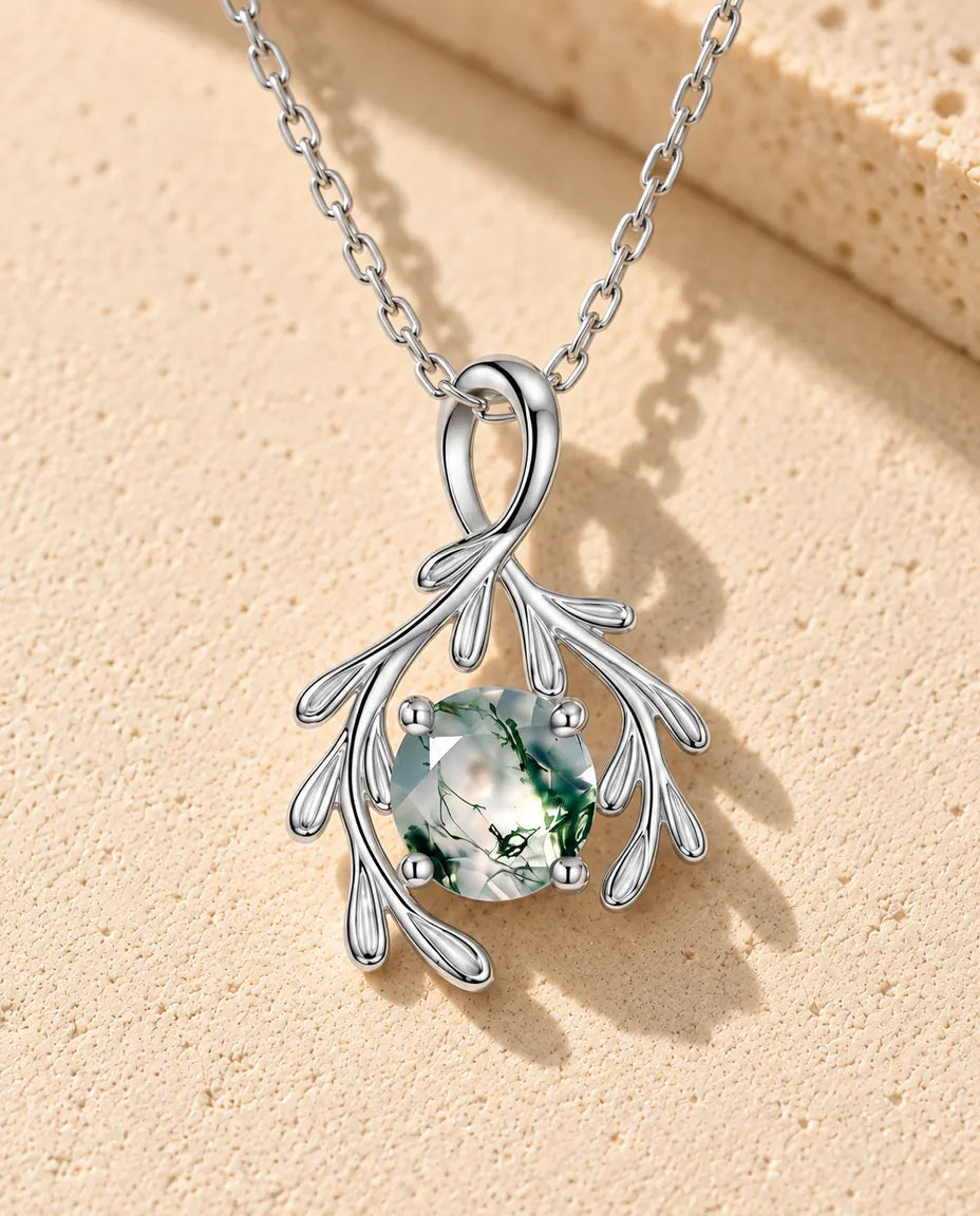 Halskæde i sølv med en grøn rund moss agate mellem to sølvgrene med små runde blade på beige puds foran travertin.