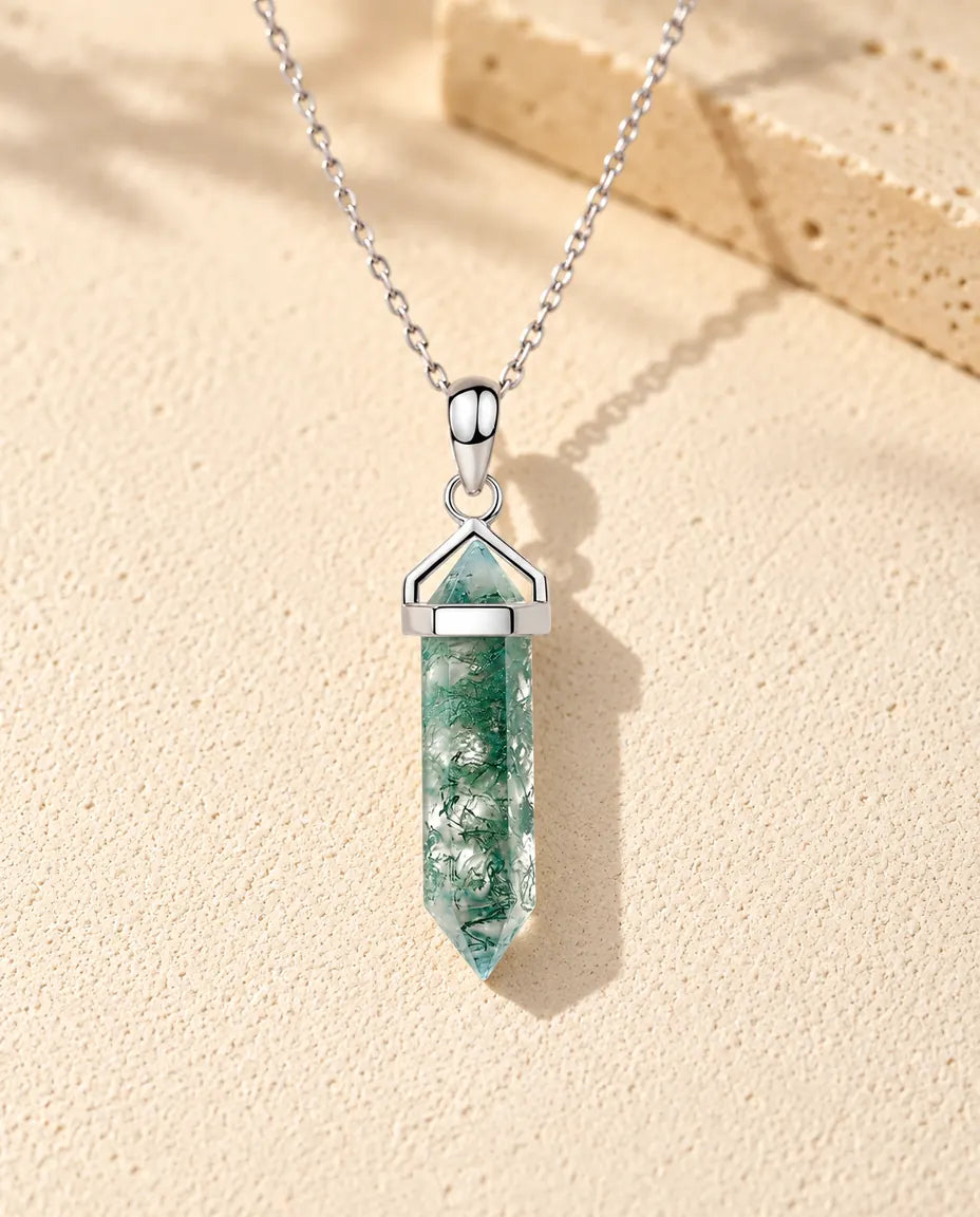 Halskæde i sølv med en lang, sekskantet grøn moss agate-spids under en sølvhætte på beige puds foran travertin.