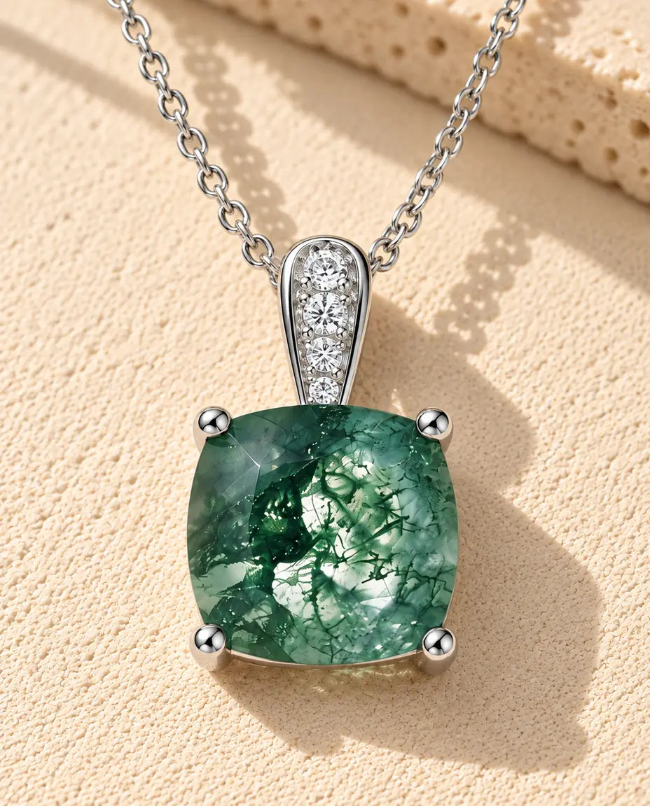 Halskæde i sølv med en grøn pudeformet moss agate i fire kløer under en øsken med små zirkonia på beige puds foran travertin.