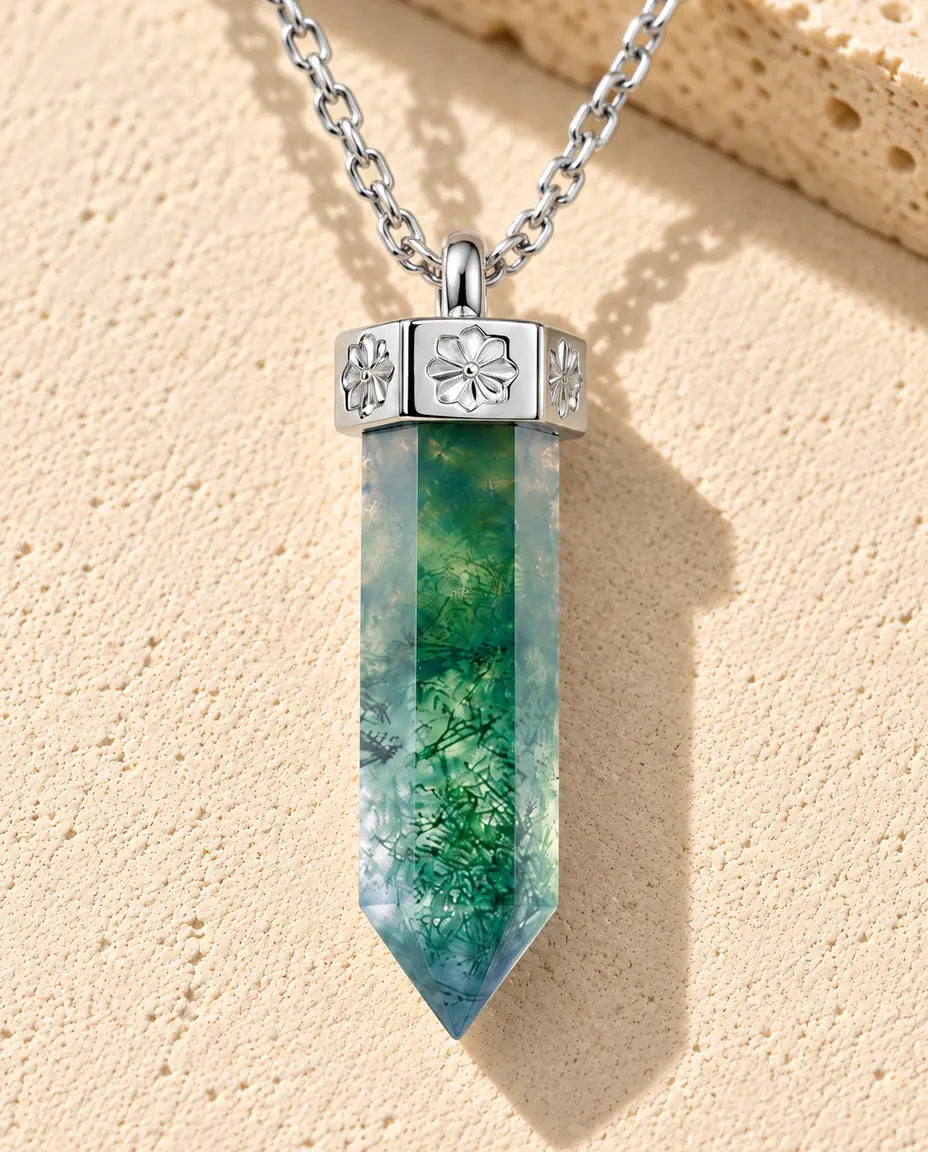 Sølvhalskæde med en grøn sekskantet moss agate-spids under en sølvhætte med små blomster på beige puds foran travertin.