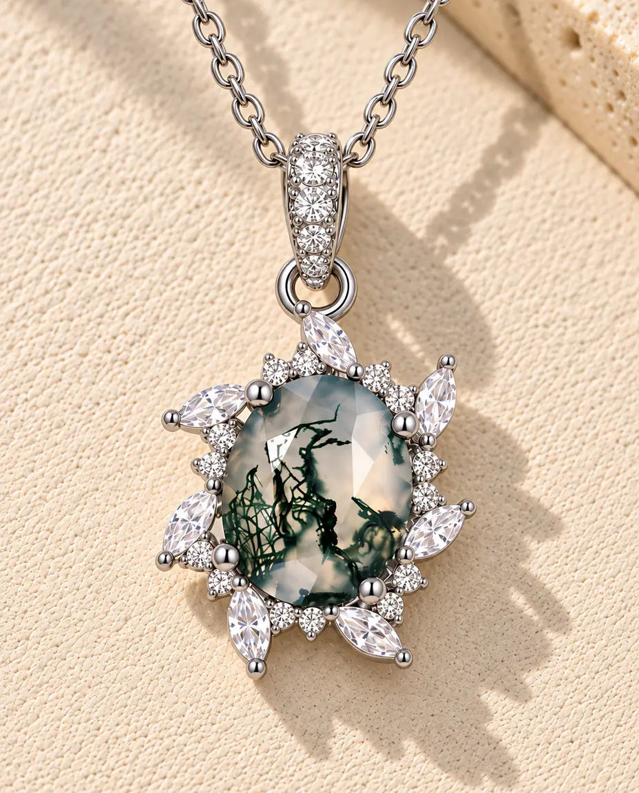 Halskæde i sølv med en grøn oval moss agate omgivet af stråler af små marquise-zirkonia på beige puds foran travertin.