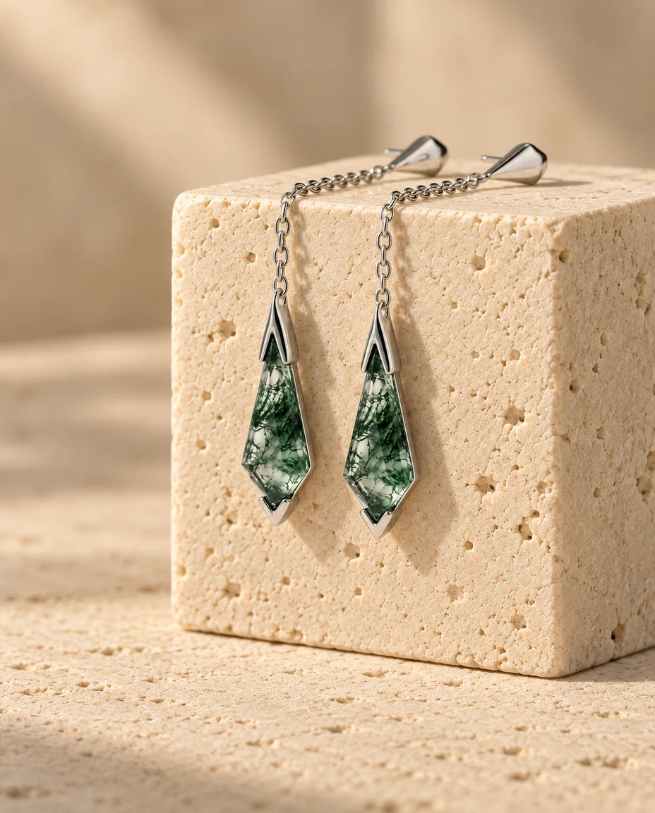 Et par øreringe i 925 sterling sølv med fin kæde og kite-slebet grøn moss agate, der hænger ned ad en beige travertinblok.