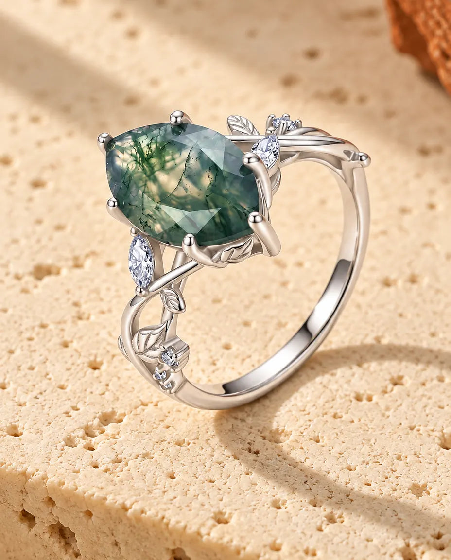 Sølvring med en facetteret Moss Agate i marquiseslibning på snoede ranker med små blade, stående på lys travertin.