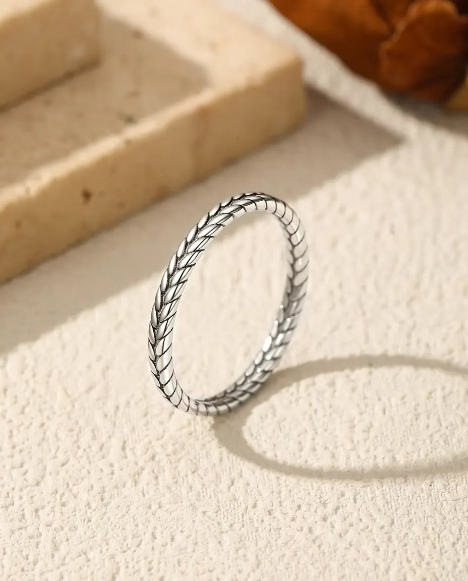 Tynd ring i 925 sterling sølv med flettet hvedeaksmønster står på groft puds med oval skygge ved travertin og tørret blomst.