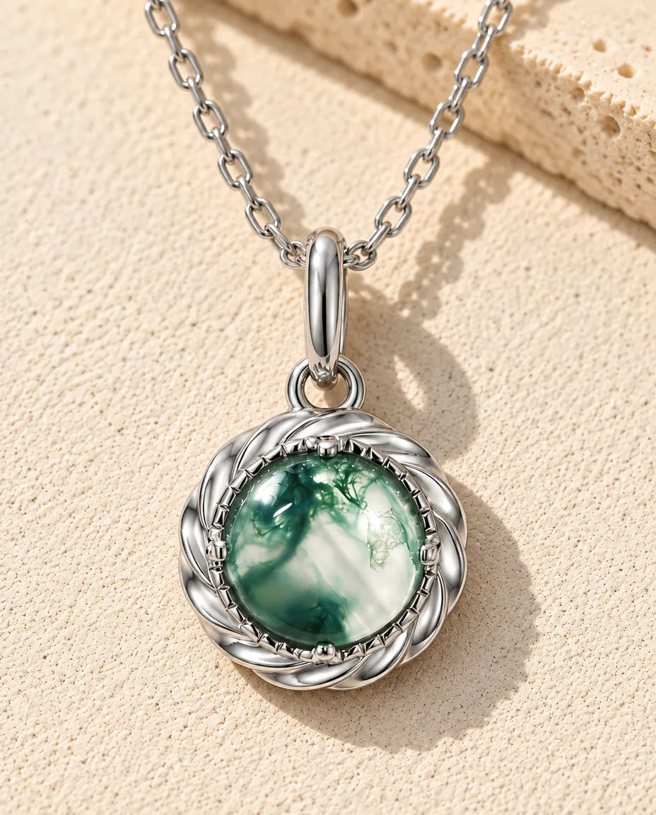 Halskæde i sølv med en rund grøn moss agate i en snoet sølvramme på beige puds foran travertin i varmt sidelys.