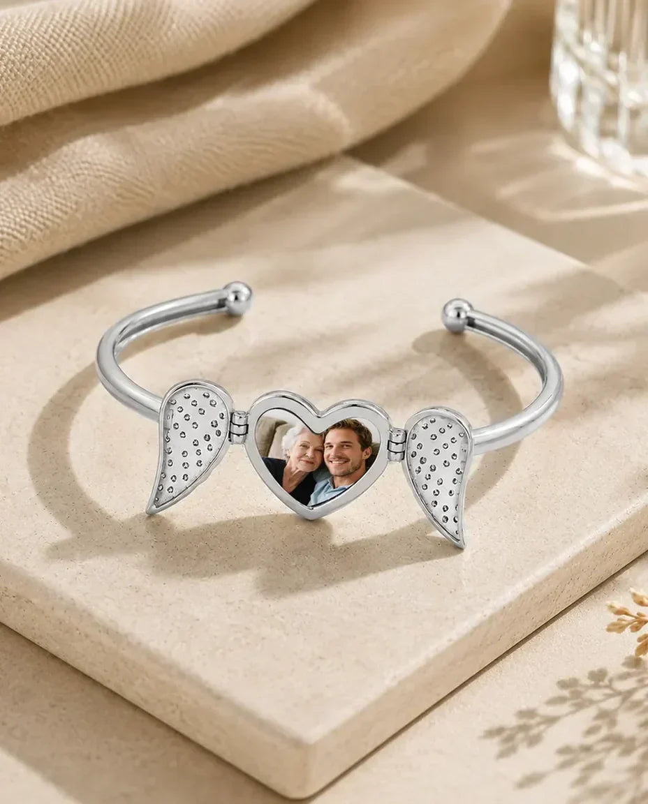 Blank bangle med en hjertemedaljon mellem to englevinger med krystaller, der åbner sig om et billede.