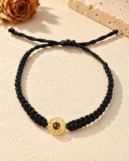 Armbånd med guldfarvet rund charm på sort flettet snor set ovenfra med glideknude og snorender