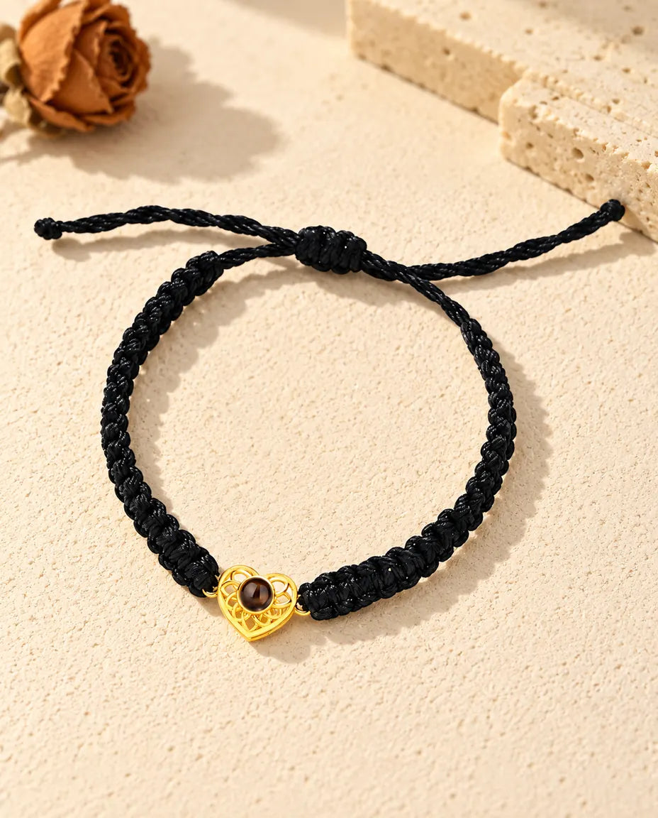 Armbånd med guldfarvet hjerte på sort flettet snor set ovenfra med glideknude og snorender