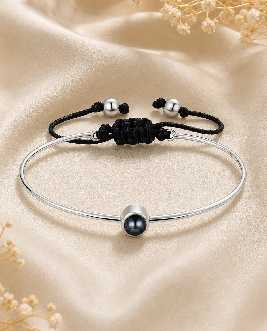 Tynd, blank bangle i sølvfarve med en lille sort perle foran og en flettet sort snor med løbeknude.
