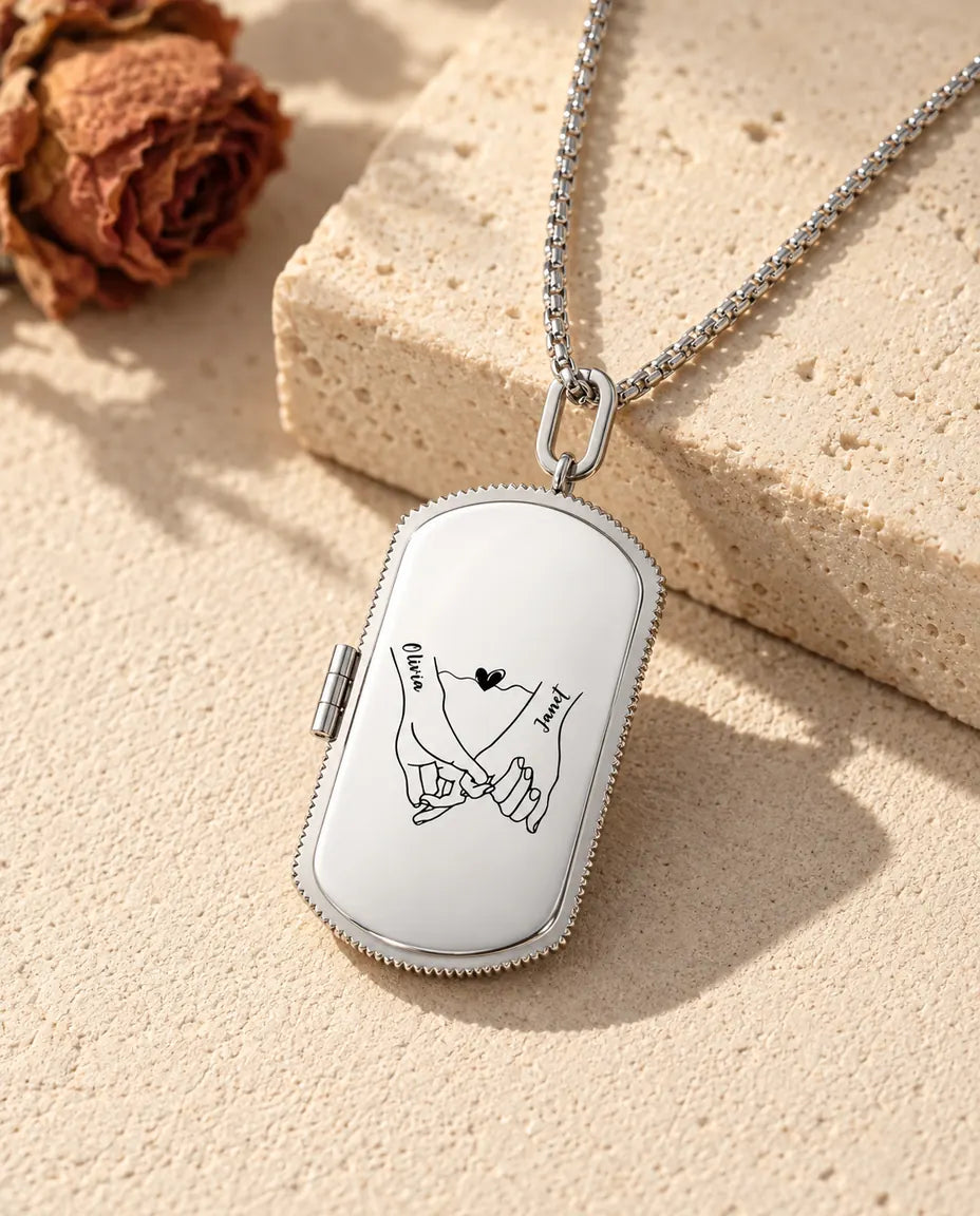 Sølvfarvet dog tag-medaljon i rustfrit stål med indgraverede hænder og to navne på en lys stenflade ved tørrede roser