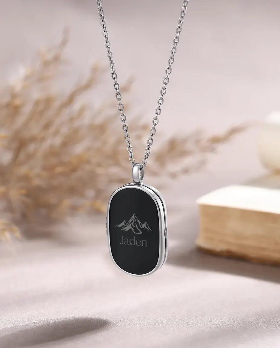 Aflang oval medaljon med mat sort forside, indgraveret bjergmotiv og navn, båret i en kæde i sølvtone.