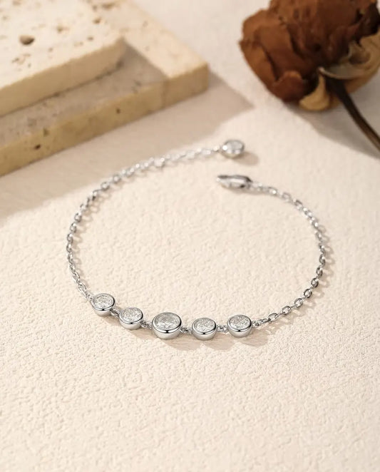 Fint armbånd i 925 sterling sølv med fem runde moissanite-sten i lukket fatning på række, lagt på råhvid puds.