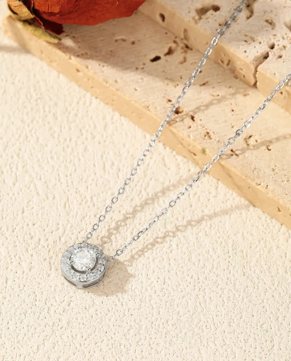 Rundt vedhæng med en moissanite i midten og en krans af små pavésten i rhodineret 925 sterling sølv.