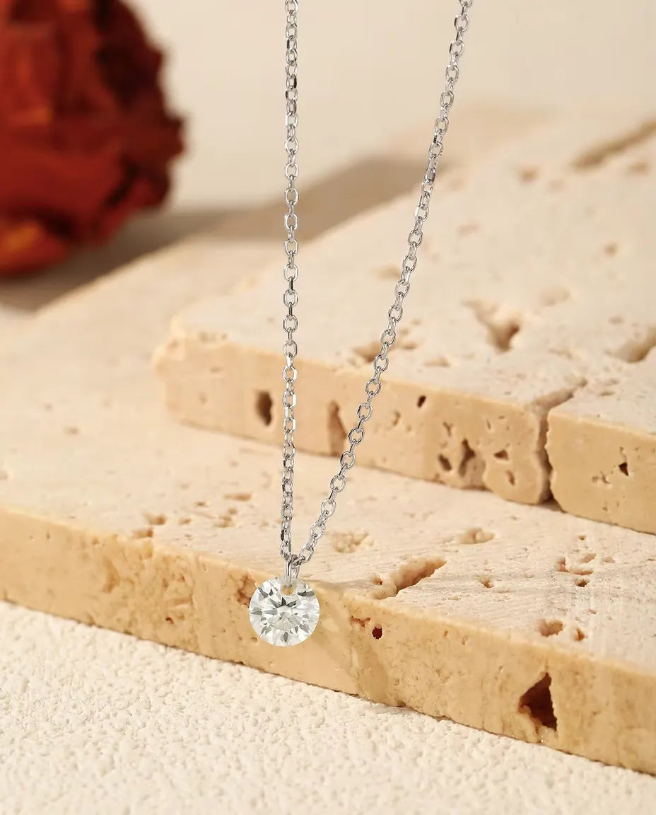 Sølvhalskæde med en rund moissanite i brillantslibning, der hænger over en lys travertinblok ved siden af en tørret rose.