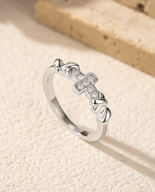 Sølvring med et vandret kors fuld af små moissanite-sten og to blanke knuder på hver side, skråt på råhvid puds.