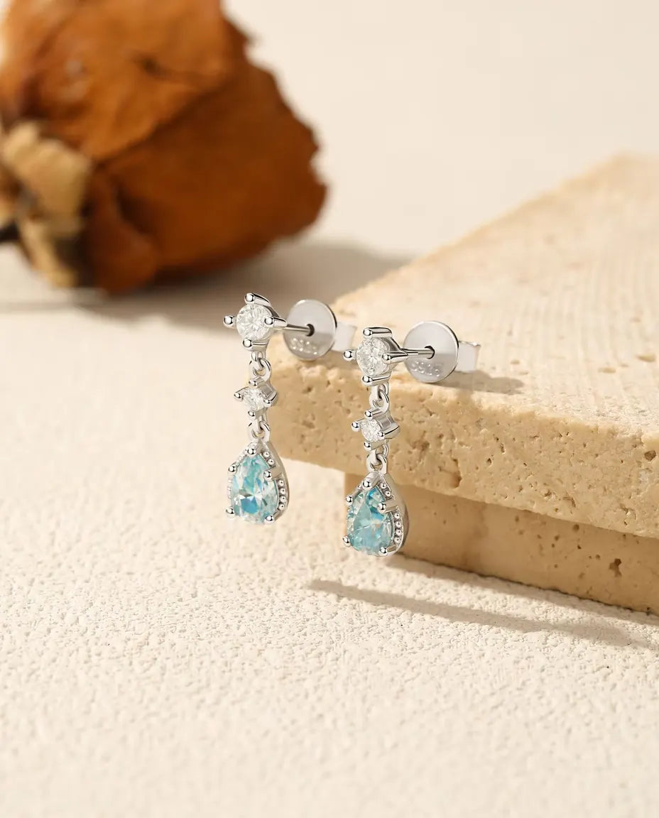 Sølvøreringe med aquablå pæreslebne moissanite i perlet ramme under to klare runde accentsten.