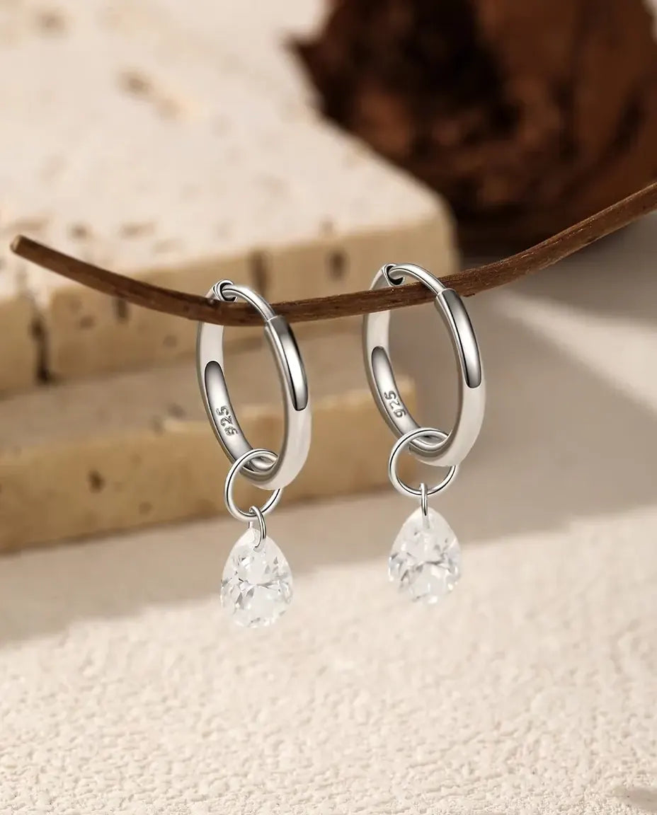 Et par glatte creoler i 925 sterling sølv med en hængende dråbeformet moissanite, hængt på en mørkebrun kvist ved travertin.