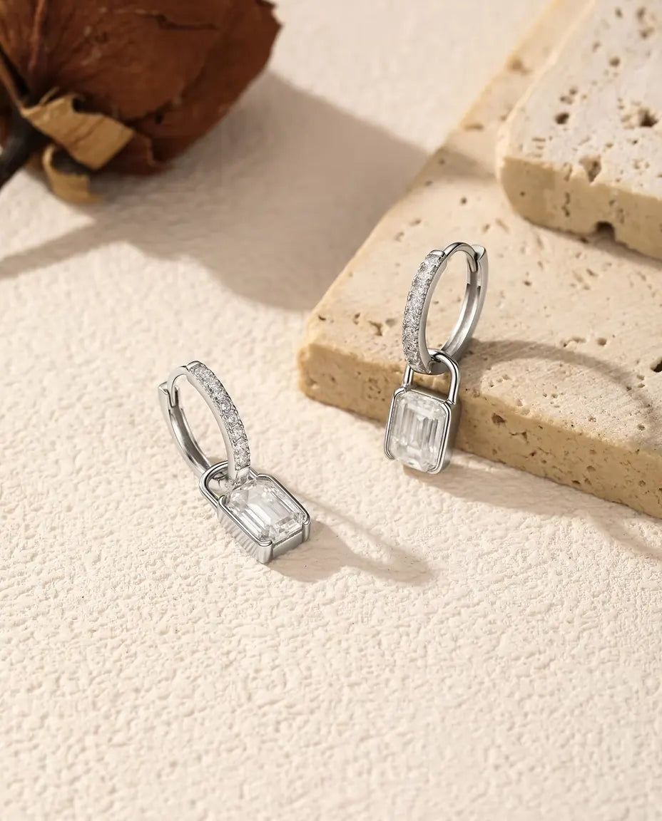 Et par creoler i 925 sterling sølv med små sten og en hængelås af moissanite, den ene lænet op ad en lys travertinblok.