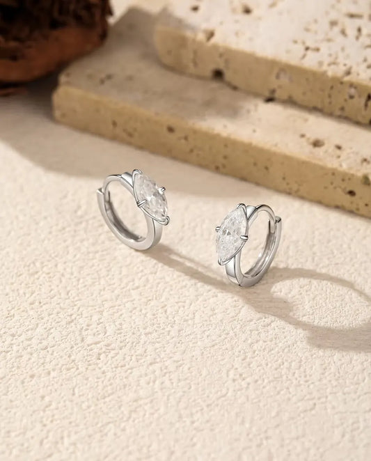 Et par små creoler i 925 sterling sølv med en marquiseslebet moissanite på tværs, stillet op på råhvid puds ved travertin.