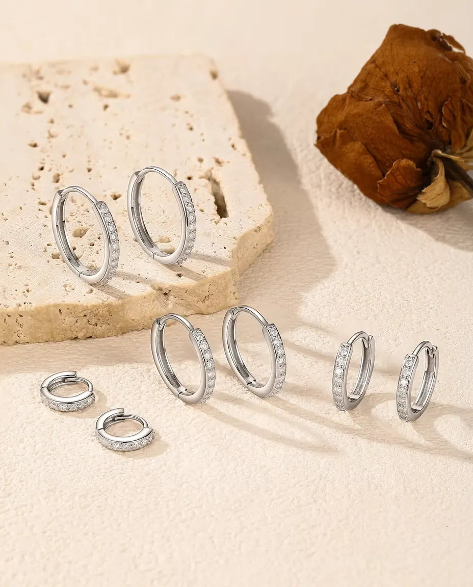 Fem par små creoler i 925 sterling sølv med pavéfattet moissanite på råhvid puds og lyse travertintrin ved en tørret rose.