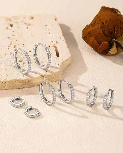 Fem par små creoler i 925 sterling sølv med pavéfattet moissanite på råhvid puds og lyse travertintrin ved en tørret rose.