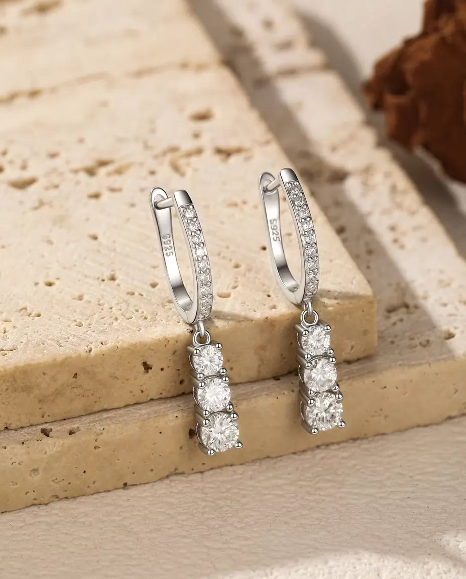 Et par små creoler i 925 sterling sølv med tre runde moissanite-sten i voksende størrelse, hængt på et lyst travertintrin.