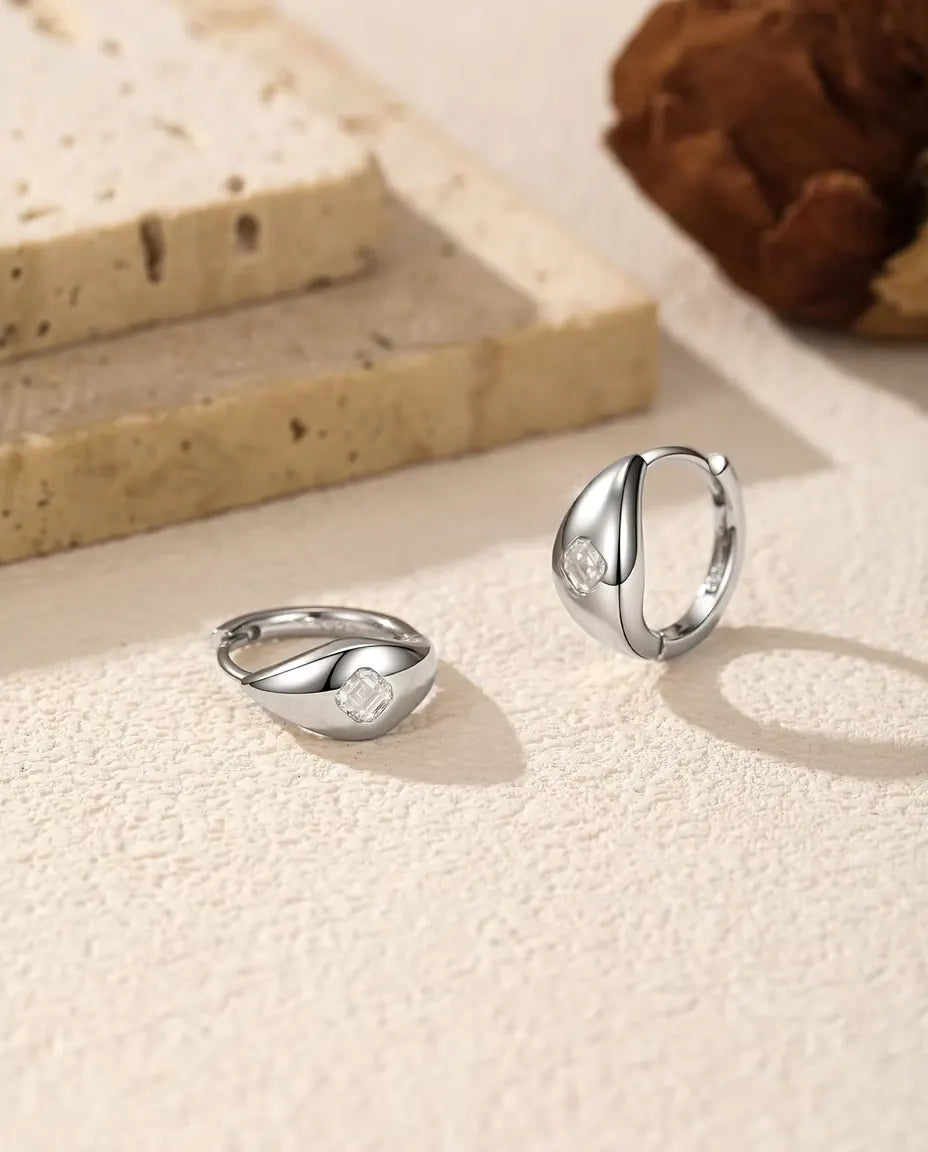 Et par små kuppelformede creoler i 925 sterling sølv med en firkantet moissanite foran, liggende og stående på råhvid puds.