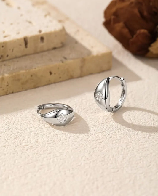 Et par små kuppelformede creoler i 925 sterling sølv med en firkantet moissanite foran, liggende og stående på råhvid puds.