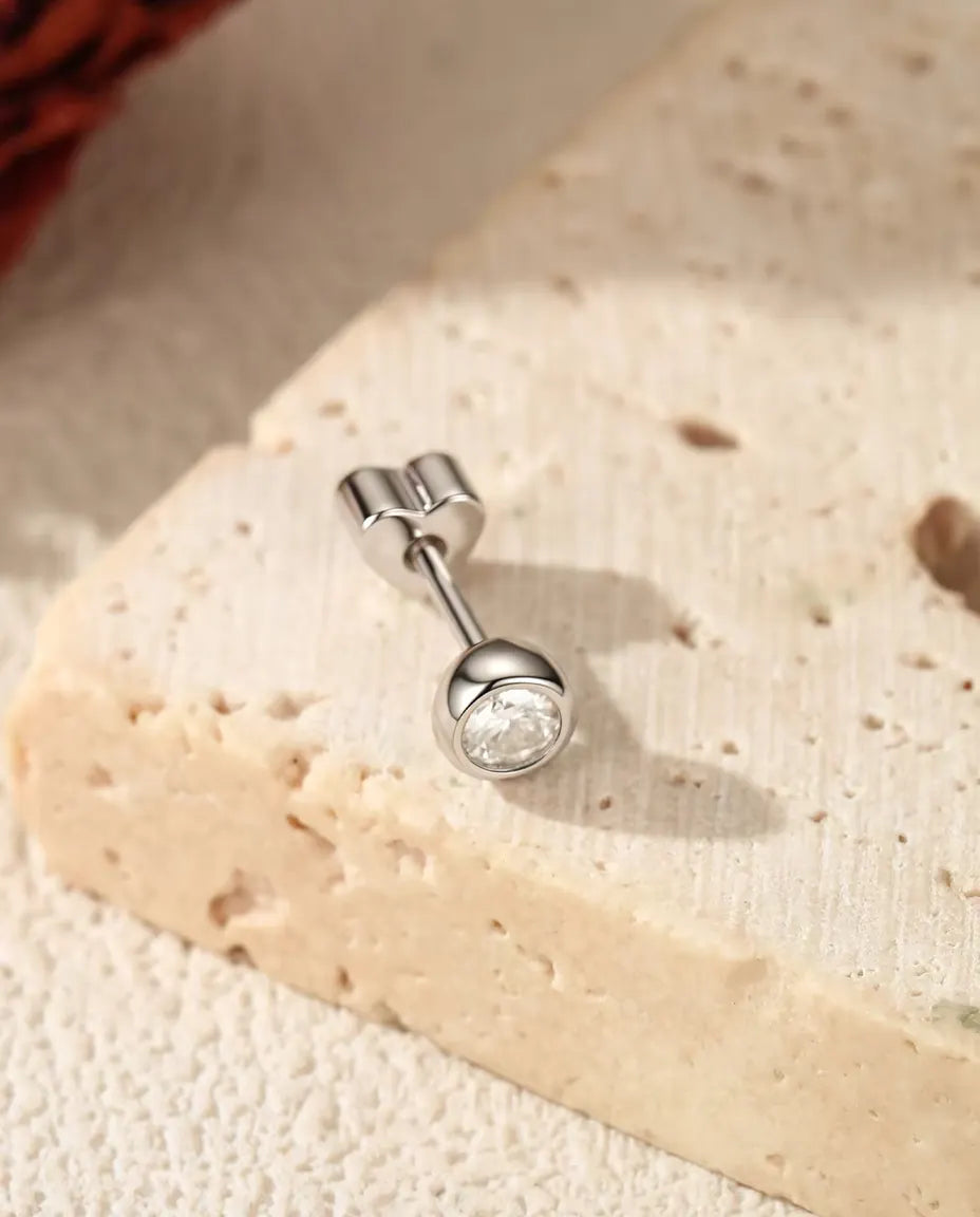 En ørestik i 925 sterling sølv med en rund moissanite på 3 mm i kantfatning, lagt på råhvid puds ved travertin.