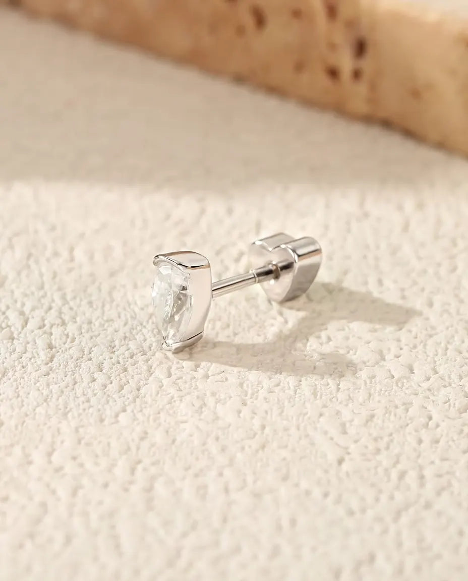 En dråbeformet ørestik med moissanite i 925 sterling sølv ligger på siden på råhvid puds, med den hjerteformede lås bagud.