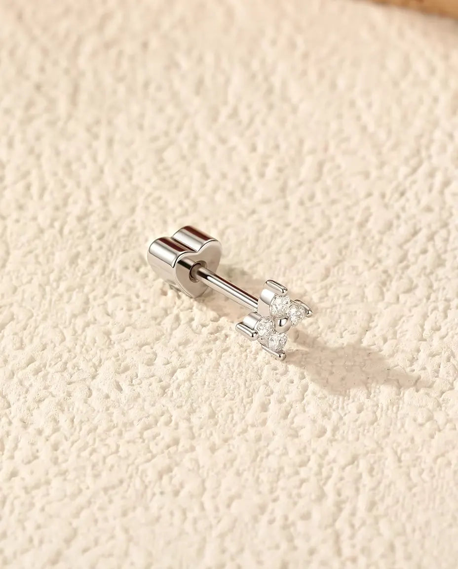En firkløverformet ørestik med moissanite i 925 sterling sølv og hjerteformet lås ligger på råhvid puds i varmt sidelys.