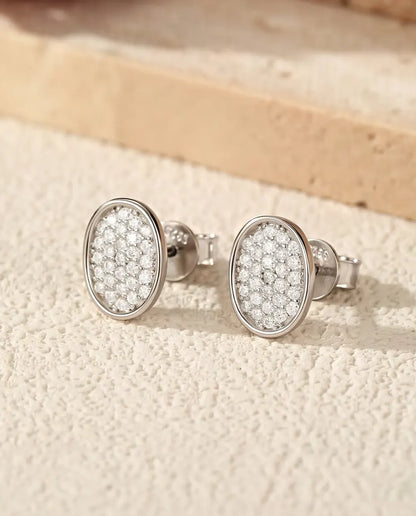 Et par ovale ørestikker i 925 sterling sølv, dækket af små moissanite-sten, står side om side på råhvid puds ved travertin.