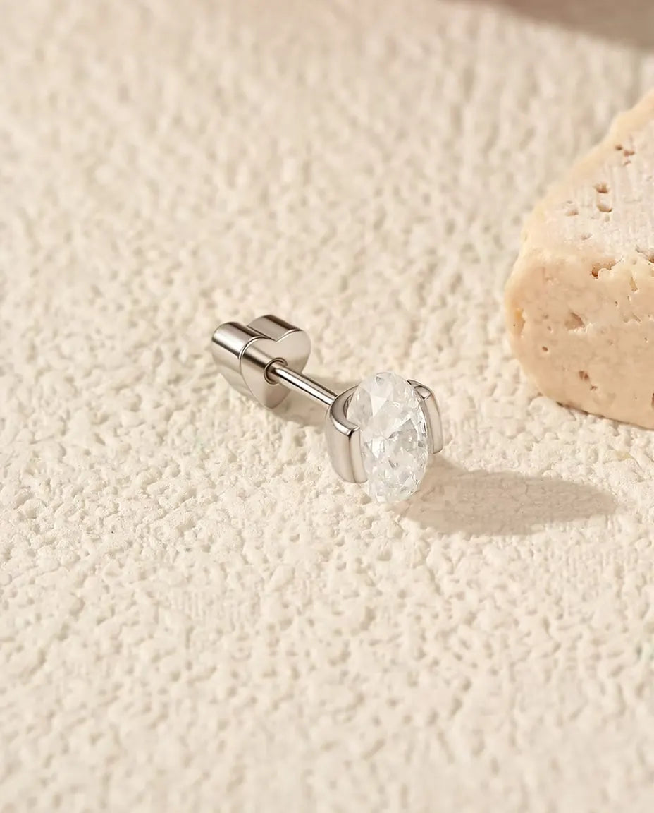 En oval moissanite-ørestik i 925 sterling sølv ligger med hjerteformet lås på råhvid puds, travertin bag til højre.