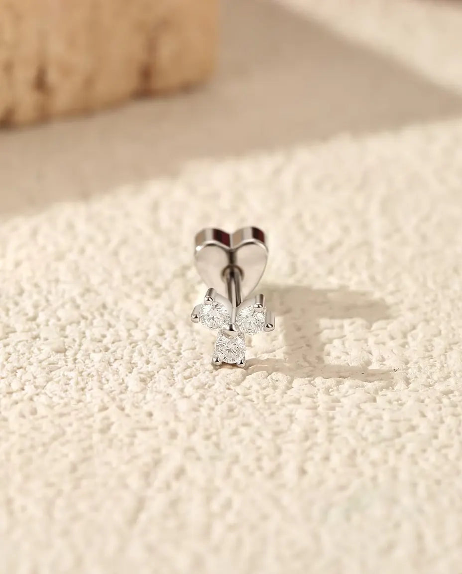 En trekløverformet ørestik med moissanite i 925 sterling sølv og hjerteformet lås står på råhvid puds foran lys travertin.